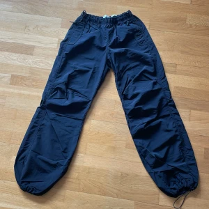 Svarta parachute pants från Bershka - Säljer ett par svarta parachute pants från Bershka med elastisk midja och dragsko i benslut. Byxorna har en loose passform och är tillverkade i ett lätt syntetmaterial som ger en cool streetkänsla. Perfekta för dig som gillar avslappnad stil.