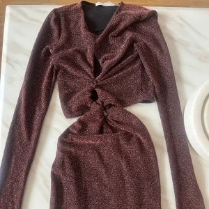 Glittrig vinröd långärmad klänning - Snygg vinröd klänning med glittrigt tyg och lång ärm. Klänningen har två twistade cutouts framtill som ger en cool och trendig look. Perfekt för dig som vill sticka ut med en unik detalj och gillar att glänsa.