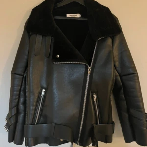 Svart teddyjacka från Chammas - Riktigt snygg svart teddyjacka från Chammas med bred krage i fuskpäls, dragkedja i silver och coola spänndetaljer på ärmar och midja. Jackan har två snedställda fickor med dragkedja och är fodrad för extra värme. Perfekt statement-plagg för kalla dagar.