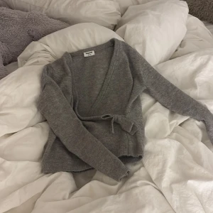 Grå omlottkofta från Kappahl - Mysig grå omlottkofta från Kappahl med knytband i midjan och långa ärmar. Koftan är stickad och har en enkel, stilren look som passar perfekt till lager-på-lager. Superskön att slänga på sig när det är kyligt ute.