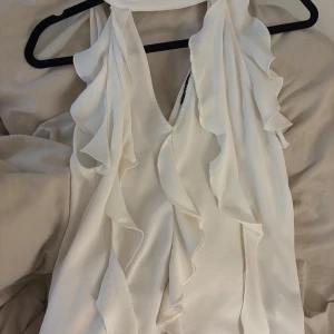 Vit med volangdetaljer - Säljer en vit blus med volanger och djup v-ringning. Blusen är ärmlös och har en snygg scarf-detalj runt halsen 