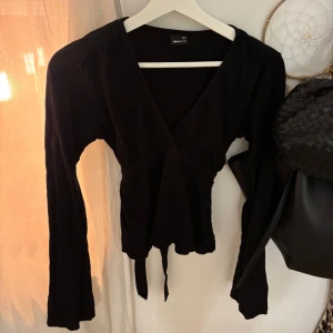 Svart topp med knyt bak från Gina Tricot - Svart topp från Gina Tricot med långa ärmar och v-ringning. Toppen har knytband i midjan som ger en snygg siluett och lätt volangkant nedtill. Perfekt att styla med jeans eller kjol för en trendig look.