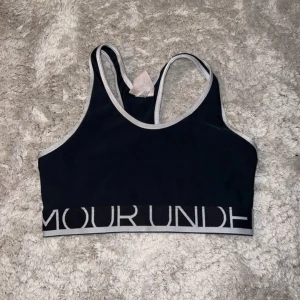 Svart sporttopp från Under Armour - Svart sporttopp från Under Armour med  kanter och bred resår med logga runt om. Snygg racerback-modell som ger bra stöd och passar perfekt till träning. Tillverkad i stretchigt syntetmaterial för maximal komfort.