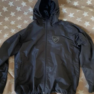 Svart vindjacka med DIF-logga - Svart vindjacka med huva och dragkedja framtill. Jackan har två sidofickor och en vit DIF-logga på bröstet. Perfekt för blåsiga dagar och enkel att matcha med det mesta. Resår i nederkant och ärmslut för skön passform.