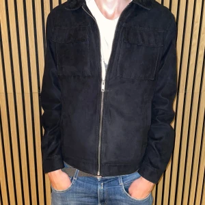 Svart mockajacka från Jack & Jones - Snygg svart mockajacka från Jack & Jones med klassisk krage och två bröstfickor med lock. Jackan har dragkedja framtill och är helfodrad. Perfekt för dig som vill ha en stilren och enkel look med lite edge.