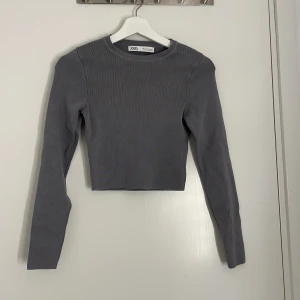 Grå croppad topp från Zara - Säljer en stilren grå croppad topp från Zara. Toppen är ribbad, har lång ärm och rund halsringning. Perfekt för dig som gillar en enkel men trendig look. Materialet är mjukt och stretchigt för en bekväm passform.
