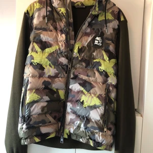 Camouflage pufferjacka från Etro - Pufferjacka från Etro med camouflage-mönster i grönt, brunt och beige samt inslag av gult. Jackan har stickade ärmar i olivgrönt, huva och två dragkedjefickor framtill. Ursprungspris 9400 kr.