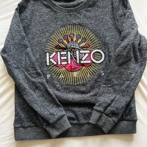 Grå Kenzo sweatshirt med brodyr - Säljer en grå sweatshirt från Kenzo med stort broderat färgglatt motiv och logga på bröstet. Tröjan har rund hals, långa ärmar och ribbade muddar. Perfekt statement piece för dig som gillar streetstyle och vill sticka ut.