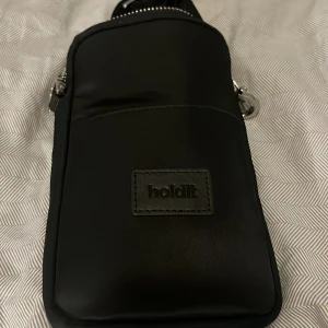 Svart axelväska från Holdit - Snygg svart axelväska från Holdit med justerbar bred axelrem med logga. Väskan har ett stilrent, minimalistiskt utseende och är tillverkad i slätt syntetmaterial. Praktisk dragkedja upptill och silverfärgade metalldetaljer. Perfekt för mobilen och småprylar.