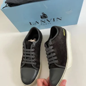 Svarta sneakers från Lanvin i mocka - Snygga svarta sneakers från Lanvin med ovandel i mocka och tåparti i grått skinn. Skorna har grå snörning, vit sula och en gul detalj på sidan. Perfekta för dig som gillar stilrena och exklusiva sneakers med en twist.