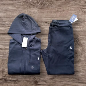 Navy blue tracksuit från Polo Ralph Lauren - Snygg mörkblå hoodie och byxor från Polo Ralph Lauren med klassisk vit logotyp på bröstet. Tröjan har huva, dragkedja framtill och två fickor. Materialet är mjuk bomull och passformen är normal. Perfekt för kyliga dagar eller när du vill ha en stilren look.