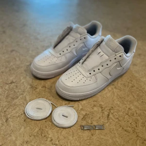 Nike Air Force 1 helvita sneakers - Klassiska Nike Air Force 1 sneakers i helvitt skinn med perforerad tå och vit sula. Snygg och tidlös modell med rund tå, platt sula och medföljande vita skosnören samt silvriga dubraes. Perfekta för dig som gillar cleana och stilrena sneakers.