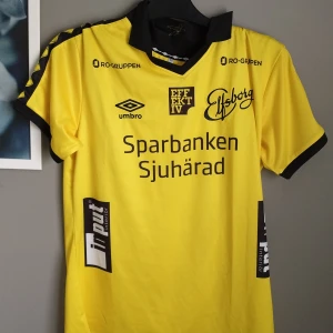Gul Elfsborg fotbollströja Umbro - Elfsborgs officiella matchtröja från Umbro i stark gul färg med svarta detaljer och tryck. Kortärmad modell med svart krage och loggor på bröstet, ryggen och ärmarna. Tillverkad i lätt polyester som andas, perfekt för träning eller match.