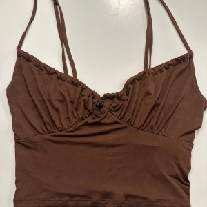 Brun croppad linnetopp från Bik Bok - Snygg brun croppad linnetopp från Bik Bok med smala axelband och rynkade detaljer vid bysten. Tillverkad i mjuk och stretchig polyesterblandning som sitter skönt på kroppen. Perfekt för en trendig och avslappnad look.