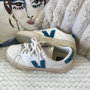 Vita Veja sneakers med blå detaljer - Säljer ett par klassiska Veja sneakers i vitt skinn med blått V på sidan och blå detalj vid hälen. Skorna har rund tå, platt sula och snörning framtill. Kan lätt rengöras lätt då det finns lite smuts på sulan osv.. jag använder oftast 37,5/38💗 