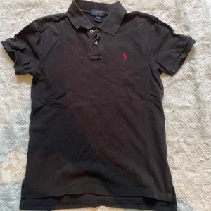 Svart Polo Ralph Lauren t-shirt (14-16y) - Svart t-shirt från Polo Ralph Lauren med klassisk pikékrage och två knappar vid halsen. Den har korta ärmar och en röd broderad logga på bröstet. Materialet är mjuk bomull och passformen är normal, perfekt för en chill och stilren look. Storleken är juniorstorlek.