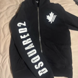 Svart hoodie från Dsquared2 med tryck - Svart hoodie från Dsquared2 med stor vit logga på ärmen och ett snyggt lövtryck på bröstet. Tröjan har dragkedja framtill, huva och två fickor. Perfekt för dig som gillar streetstyle och vill sticka ut med ett coolt märke. Storlek 14y sitter som s/xs
