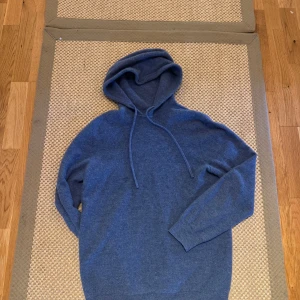 Blå hoodie i kashmir med huva - Mysig blå hoodie i merinoull med huva och snörning. Tröjan har långärmad design och ribbade muddar vid ärmslut och nederkant. Perfekt för dig som vill ha en stilren och bekväm look.