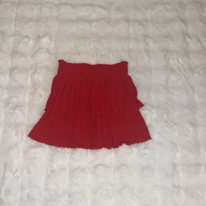 Röd volangkjol med resårmidja - Supersöt röd kortkjol med volanger och smockad resår i midjan. Kjolen har en flowy känsla och passar perfekt till sneakers eller boots. Enkel att styla med en crop top eller stickad tröja.