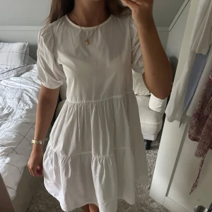 Vit kort klänning med puffärm - Supersöt vit klänning med rund hals och puffiga korta ärmar. Modellen är kort med volanger i kjolen som ger en flowy och chill vibe. Perfekt för varma dagar och enkel att styla med smycken. Materialet känns lätt och mjukt mot huden.