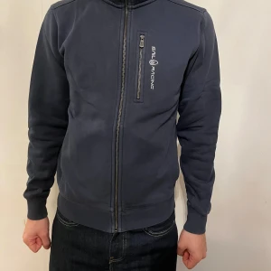 Mörkblå hoodie från Sail Racing - Mörkblå hoodie från Sail Racing med dragkedja framtill och en bröstficka med logga. Tröjan har huva, långa ärmar och ribbade muddar. Perfekt för dig som gillar en sportig och clean stil.