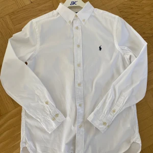 Vit skjorta från Polo Ralph Lauren - Klassisk vit skjorta från Polo Ralph Lauren i slim fit-modell. Skjortan har button down-krage, långa ärmar och broderad marinblå logga på bröstet. Tillverkad i bomull och stängs med vita knappar framtill. Perfekt för en clean och stilren look.