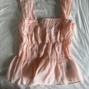 Ljusrosa volangig linnetopp XS - Supersöt ljusrosa linnetopp med breda axelband och volangig nederdel. Smockad över byst och midja för en skön passform. Tunt, luftigt tyg som är perfekt för varma dagar. Enkel och romantisk vibe, passar till jeans eller kjol.