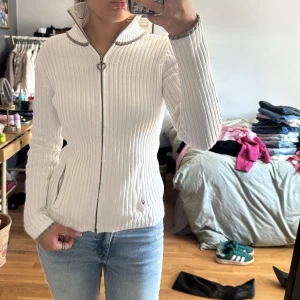 Vit ribbad zip up kofta  - Vit ribbstickad kofta i 100% bomull. Långärmad med dragkedja hela vägen och snygga kontrastdetaljer i grått vid krage och ärmslut. 