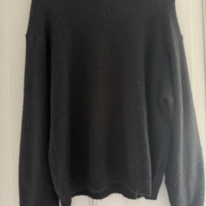 Svart stickad tröja med rund hals - En svart stickad tröja med rund halsringning och långa ärmar. Tröjan har ribbade muddar vid ärmslut och nederkant, vilket ger en klassisk och enkel look. Perfekt för lager-på-lager under kyliga dagar.