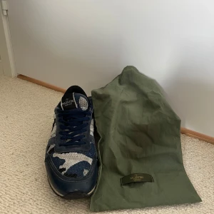 Valentino Garavani Rockrunner sneakers - Säljer ett par Valentino Garavani Rockrunner sneakers i blått och grått med coolt camouflage-mönster. Skorna har snörning, platt sula och detaljer i mocka och textil. Kommer med original dustbag och äkthetskort. Perfekta för dig som gillar lyxiga och unika sneakers.