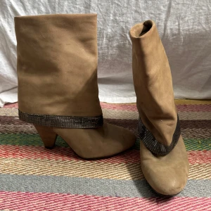 Guess boots med klack och metallicdetalj - Snygga beiga boots i mocka med bred skaft och markerad klack. Runt skaftet sitter ett band med metallicskimrande rutmönster som ger extra edge. Perfekta för dig som vill sticka ut med en cool retrostil.