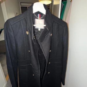 Svart rock från Tommy Hilfiger - Stilren svart rock från Tommy Hilfiger med snygga guldknappar och klassisk krage. Jackan har långa ärmar och två fickor framtill. Perfekt för dig som vill ha en tidlös och elegant look.