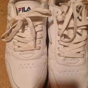 Vita sneakers från FILA, storlek 40 - Klassiska vita sneakers från FILA med broderad logga i blått och rött på sidan och hälen. Skorna har rund tå, snörning och är tillverkade i syntetmaterial med textilfoder. Perfekta för en clean och sportig stil. Knappt använda. Snöret lite smutsigt på ena skon, se bild. 