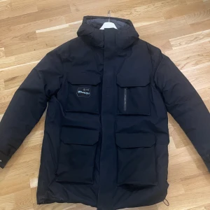 Svart dunjacka med stora fickor - Snygg svart dunjacka från sail racing med fyra stora fickor framtill, hög krage och huva. Jackan har dragkedja och är perfekt för kalla dagar. Stilren design som funkar till det mesta och håller dig varm hela vintern.