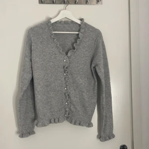 Grå stickad kofta med volangkant - Superfin grå stickad kofta från SheIn med volangdetaljer längs knäppningen, ärmslut och nederkant. Koftan har pärlknappar och lång ärm, perfekt för lager på lager. Enkel att styla till både jeans och kjol för en söt look. (Aldrig använd)