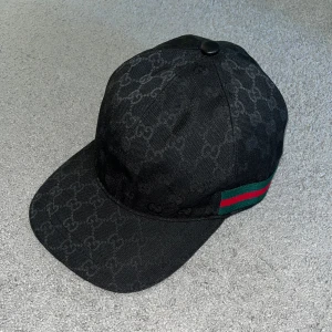 Svart Gucci keps  - Säljer en svart keps från Gucci med klassiskt GG-monogram över hela kepsen och ikoniska gröna och röda ränder på sidan. Justerbar rem baktill i skinn och snygg, böjd skärm. Materialet är slitstark bomull och polyester, perfekt för dig som gillar exklusiv streetstyle. Äkta