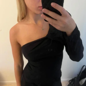 Svart topp med puffärm - Snygg svart topp med en axel och puffig lång ärm. Toppen har en figurnära passform och är tillverkad i ett glansigt material som ger en lyxig känsla. 