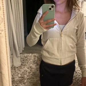 Vit hoodie Juicy Couture  - Säljer en supermjuk och lyxig vit hoodie från Juicy Couture i velour. Tröjan har guldiga detaljer på dragkedja och snören, huva och känguruficka framtill. Perfekt för en chill och trendig look.