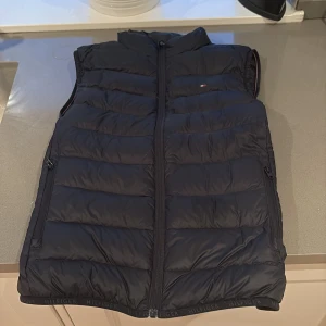 Mörkblå dunväst från Tommy Hilfiger - Snygg mörkblå dunväst från Tommy Hilfiger med quiltad design och dragkedja framtill. Västen har diskret logga på bröstet och Hilfiger-detaljer längs nederkanten. Perfekt att slänga över en hoodie för en chill look.
