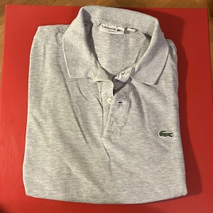 Grå långärmad pikétröja Lacoste - Snygg grå långärmad pikétröja från Lacoste med klassisk krage, knäppning framtill och den ikoniska gröna krokodilloggan på bröstet. Tröjan är i bomull och har en slim passform som sitter schysst på kroppen.