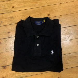 Svart pikétröja Ralph Lauren slim fit - Svart pikétröja från Ralph Lauren i modellen Custom Slim Fit. Klassisk design med vit broderad logga på bröstet, krage och två knappar framtill. Tillverkad i mjuk bomull, perfekt för en clean och stilren look.