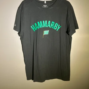 Svart Hammarby t-shirt  - Svart t-shirt från Craft med grönt Hammarby-tryck och en liten flagga på bröstet. Klassisk rund hals och korta ärmar, perfekt för dig som vill visa ditt stöd för Hammarby. Skön bomullskänsla och enkel stil.