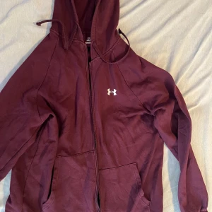 Vinröd hoodie från Under Armour - Säljer en vinröd hoodie från Under Armour med dragkedja och huva. Tröjan har en klassisk passform, långa ärmar och en vit logga på bröstet. Storlek L