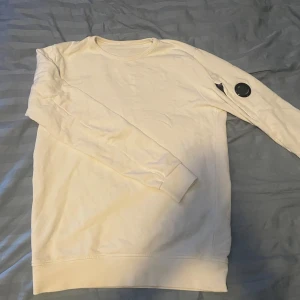 Vit sweatshirt från C.P. Company - Säljer en clean vit sweatshirt från C.P. Company i storlek S. Tröjan har lång ärm och den ikoniska linsen på vänster ärm. Tillverkad i mjuk bomull med rund hals och ribbade muddar. Perfekt för dig som gillar streetwear och stilrena detaljer.