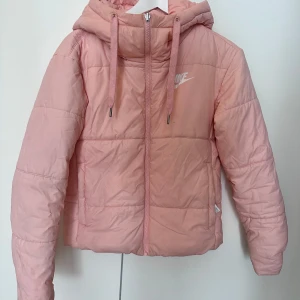 Rosa pufferjacka från Nike - Säljer en snygg rosa pufferjacka från Nike med huva och dragkedja framtill. Jackan har stora loggor både fram och bak, samt dragsko i huvan. Perfekt för kyliga dagar och riktigt skön att bära. Fodrad och med två fickor på sidorna. Som dessutom går att vända ut och in.