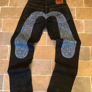 Evisu jeans - Säljer ett par mörkblå evisu jeans med unikt blått och svart abstrakt tryck på baksidan av benen. Jeansen har klassisk femficksdesign, kontrastsömmar och läderpatch i midjan. Raka ben och cool streetkänsla, perfekta för dig som vill sticka ut.
