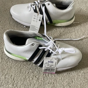 Adidas vita golfskor med Boost-sula - Snygga vita golfskor från Adidas med svarta ränder och limegröna detaljer. Skorna har en Boost-sula för extra komfort och grepp med spikar undertill. Ovandelen är i skinn och de har klassisk snörning. Perfekta för dig som vill ha stil och funktion på golfbanan. Tour 360, en av de bästa på marknaden, även Ludvig Åbergs go-to! Skorna är helt oanvända då de är för små för mig.