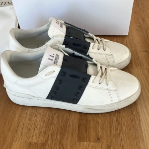 Valentino Garavani Rockstud sneakers - Snygga vita Valentino Garavani Rockstud sneakers i skinn med bred svart rem över vristen dekorerad med svarta nitar. Klassisk rund tå, vita snören och platt sula. Kommer med originalkartong och dustbags. Perfekta för dig som gillar exklusiva detaljer. Sparsamt använda lite smutsiga på sulan men bara ytligt som kan tvättas bort.