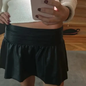 Svart läder kortkjol - Snygg svart kortkjol, rynkad midja och lätt utsvängd form. Kjolen har en stilren look och är tillverkad i ett material som ger en mjuk glans. .