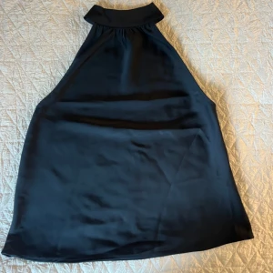 Svart halterneck topp från Bik Bok - Säljer en svart kort halterneck-topp från Bik Bok i glansigt satintyg. Toppen har hög hals, är ärmlös och stängs med dragkedja i sidan. Perfekt för dig som gillar stilrena och trendiga plagg. Endast testad, i nyskick
